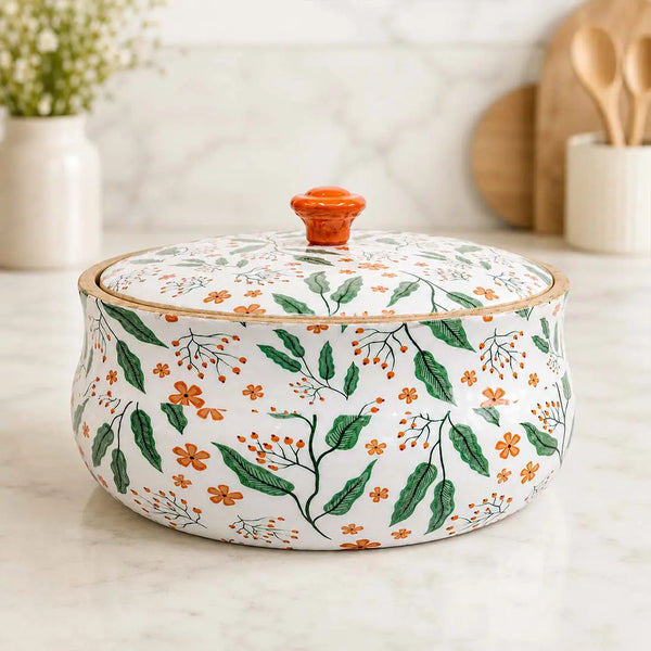 A round lidded ceramic pot with a white background and a green and orange botanical pattern, featuring a small orange knob on top, set on a light marble countertop.